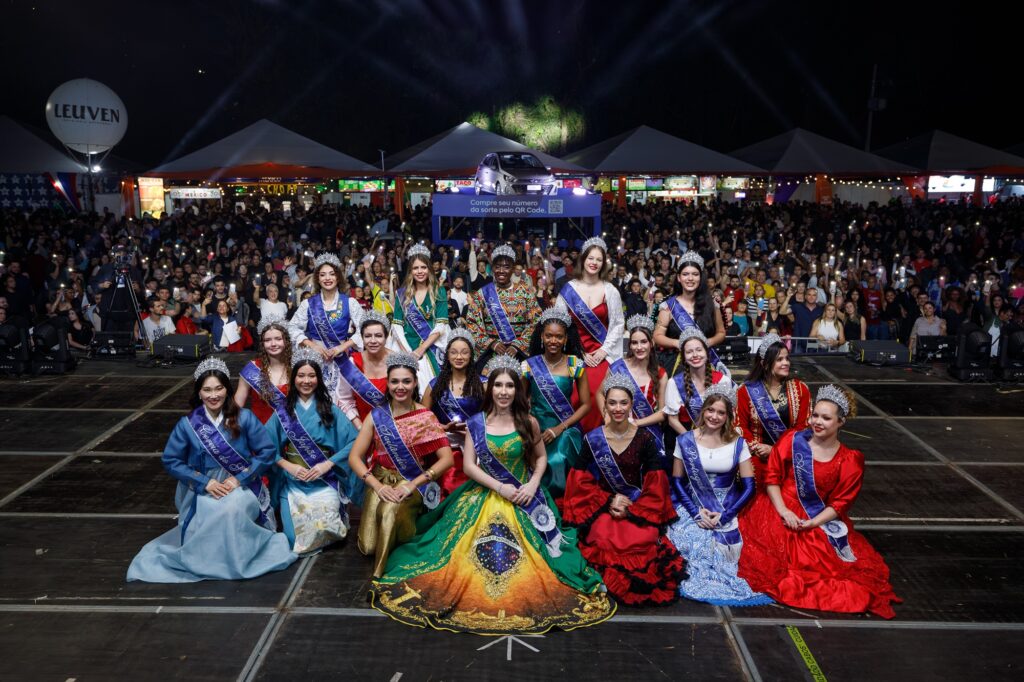 Na imagem, vemos uma cena da Festa das Nações, com um grupo de pessoas em trajes tradicionais e elaborados, cada uma usando faixa e coroa, posicionadas em um palco iluminado diante de uma grande plateia. Ao fundo, nota-se o ambiente festivo com barracas, tendas e público reunido, reforçando o clima de celebração coletiva. Acima da multidão, há uma plataforma com um carro exposto e um anúncio incentivando a compra de números da sorte via QR Code.