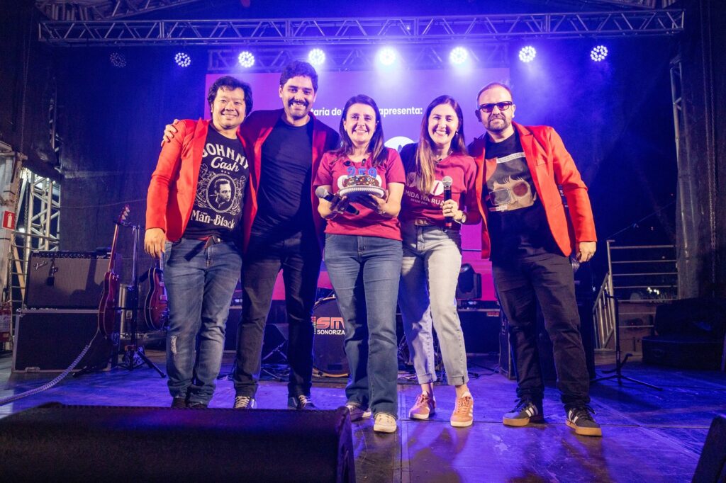 Foto de grupo num palco durante um evento noturno, com cinco pessoas alinhadas ao centro, iluminação de show e equipamento de som ao fundo. Composição: cinco pessoas lado a lado, abraçadas pelos ombros; duas mulheres ao centro e três homens (dois nas extremidades e um entre a mulher da esquerda e o homem da esquerda). Roupas e cores: três pessoas (os dois homens das extremidades e o homem do meio-esquerda) usam blazers ou jaquetas vermelhas; as duas mulheres centrais usam camisetas vinho/roxa. Jeans e tênis casuais nas pessoas visíveis. Objetos e gestos: a mulher no centro segura um pequeno bolo decorado com confeitos e velas num suporte; a mulher à direita do bolo segura um microfone; a expressão de todos é sorridente e de celebração. Palco e iluminação: grande estrutura metálica e refletores no topo iluminam em tons roxos/azuis; um telão ao fundo exibe um conteúdo parcialmente visível. Equipamentos (amplificadores, guitarras, bateria) aparecem nas laterais e atrás do grupo. Atmosfera: clima festivo e coletivo — parece comemoração ou encerramento de apresentação musical; público e som sugerem evento organizado. Detalhes menores: o bolo tem velas/numeração colorida; iluminação cria sombras e destaque nas faces; há cabos e suportes visíveis no piso do palco.