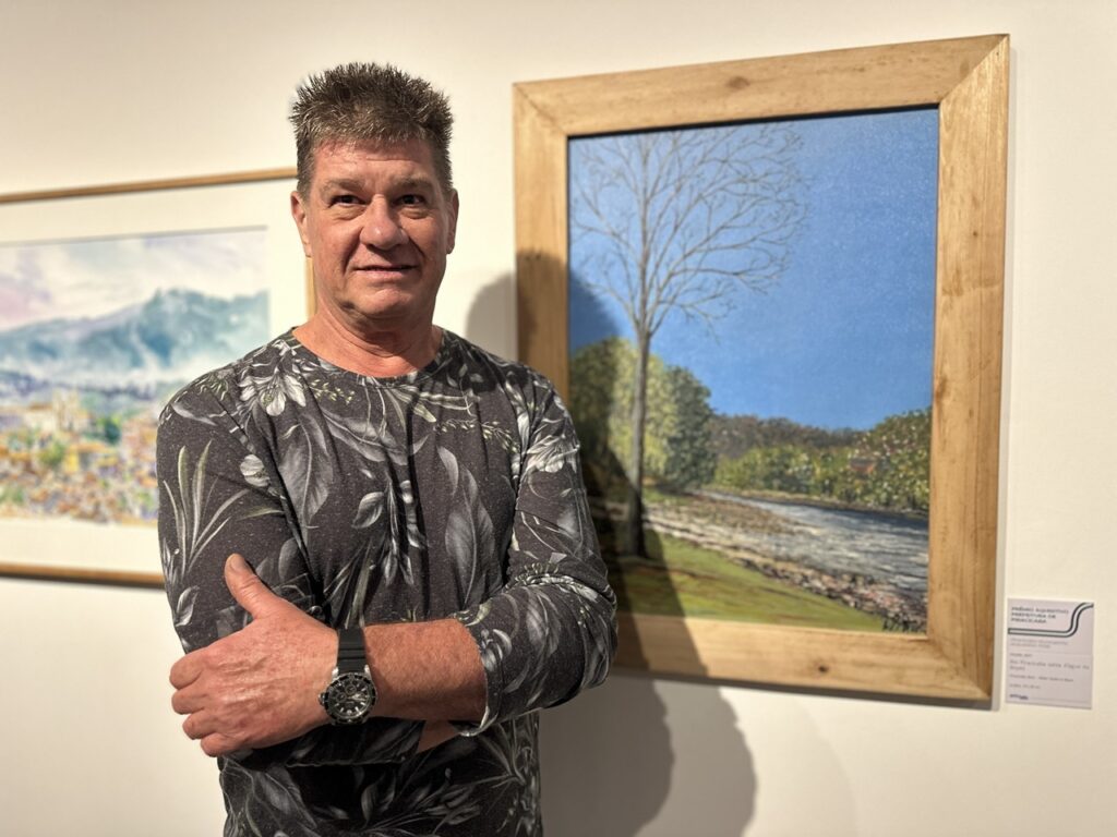 A imagem mostra um homem posando em uma galeria de arte. O homem: Ele está em primeiro plano, com os braços cruzados, vestindo uma camiseta de manga longa preta com uma estampa de folhas em tons de cinza e branco. Ele usa um relógio de pulso preto e mantém uma expressão tranquila enquanto olha diretamente para a câmera. A obra de arte: Ao lado dele, há uma pintura emoldurada em madeira clara, que retrata uma paisagem natural com uma árvore alta e esguia à esquerda, um rio e uma vegetação ao fundo sob um céu azul. Detalhes: À direita da moldura, na parede branca, vê-se uma pequena placa informativa (a ficha técnica da obra) com os dizeres "Prêmio Aquisitivo Prefeitura de Piracicaba", indicando que a peça faz parte de uma exposição institucional. À esquerda, parcialmente visível, há outra obra de arte pendurada na parede. O ambiente é iluminado e limpo, característico de espaços de exposição.