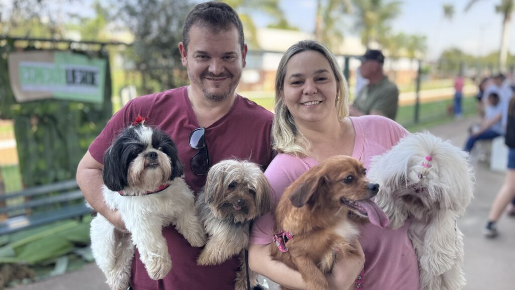 Na foto, duas pessoas estão em um ambiente ao ar livre, cada uma segurando dois cães de pequeno porte. À esquerda, alguém com camiseta vinho segura um cachorro preto e branco e outro de pelagem clara e longa. À direita, uma pessoa de camisa rosa segura um cão marrom-avermelhado com a língua de fora e um cachorro branco e peludo com um laço rosa no pelo. Ao fundo, aparecem árvores, outras pessoas e um cartaz parcialmente visível.