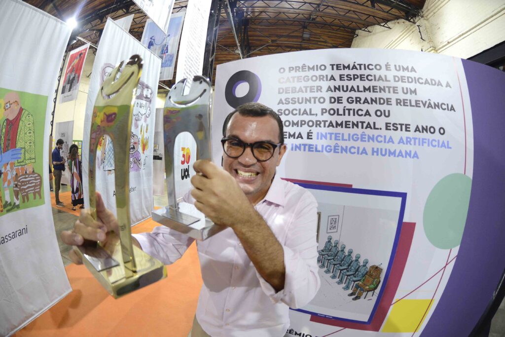 A imagem mostra um homem sorrindo e usando óculos, sentado em uma mesa durante um evento. Ele veste uma camiseta branca e segura um pincel ou marcador, aparentemente trabalhando em uma atividade artística. À frente dele há uma pintura colorida, com formas geométricas em tons de roxo, azul, vermelho, amarelo e verde. Ao fundo, várias obras de arte estão expostas em painéis, e faixas brancas pendem do teto. A foto parece ter sido tirada em uma oficina ou exposição de arte inclusiva.