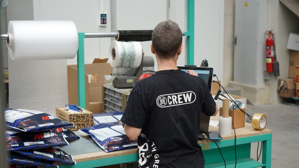A imagem mostra um trabalhador em uma área de produção, realizando atividades de embalagem e conferência de produtos em uma bancada.