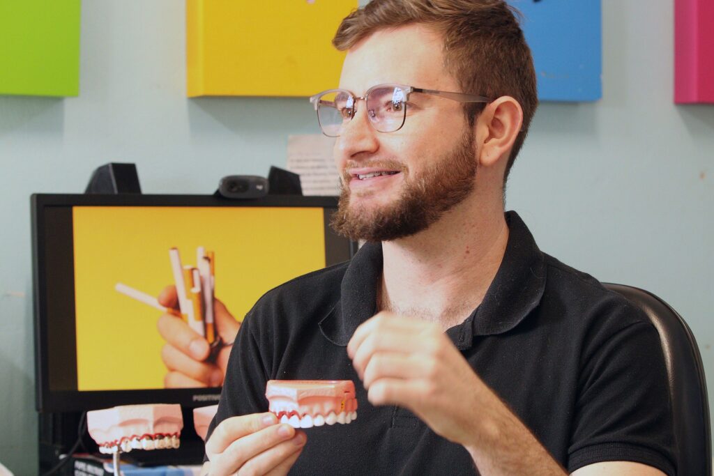 Homem adulto, sorrindo, em ambiente interno que parece ser consultório ou sala educativa. Detalhes observáveis: Aparência: cabelos curtos castanho-escuros, barba por fazer bem aparada, usa óculos com armação translúcida. Pele clara com leves sardas ou pequenas marcas no rosto. Expressão e gesto: olha levemente para a direita com expressão amistosa e atenta; sorriso mostrando os dentes; mão direita segura um modelo de arcada dentária (prótese/placa de demonstração) e a mão esquerda faz um gesto explicativo. Roupa: polo preta simples. Objetos e cenário: ao fundo há um monitor com imagem predominantemente amarela (mostrando mãos segurando objetos tipo palitos), e à esquerda dois modelos de dentes sobre suporte; painéis coloridos (verde, amarelo, azul, rosa) pendurados na parede. Mesa com materiais e possivelmente papéis atrás. Iluminação e atmosfera: luz suave e ambiente claro; sensação acolhedora e profissional — provavelmente uma demonstração ou explicação sobre saúde bucal ou próteses dentárias. Observação: descrevi apenas elementos visíveis; não faço suposições sobre identidade, profissão ou condição pessoal além do que a imagem mostra.