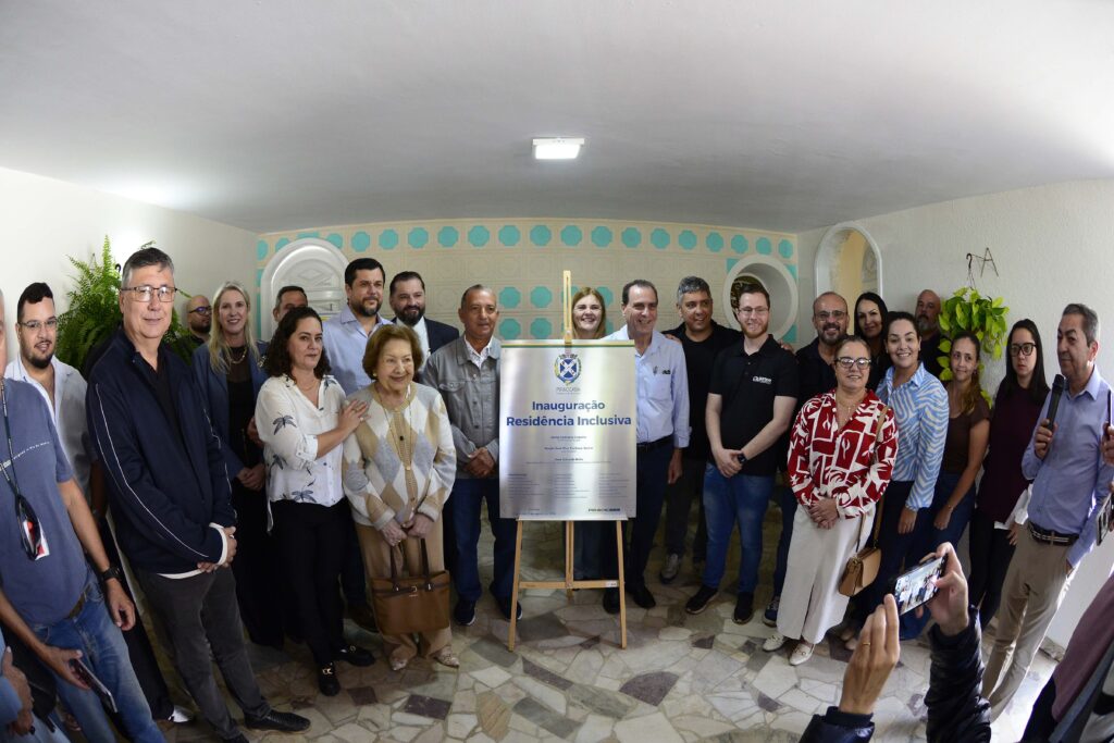 A imagem mostra um grupo de pessoas reunidas em um ambiente interno para uma inauguração oficial. No centro, há uma placa metálica sobre um cavalete de madeira com o título “Inauguração Residência Inclusiva”, indicando que o evento celebra a abertura de uma instituição voltada à inclusão social.