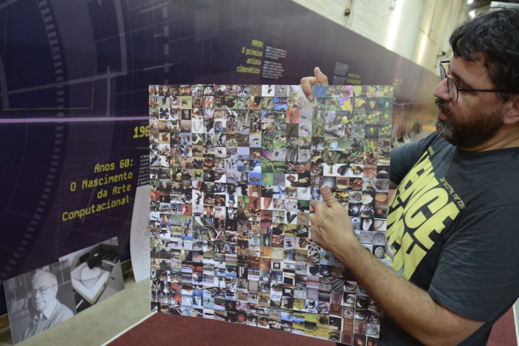 A foto mostra uma pessoa usando óculos, parcialmente visível no lado direito, segurando um grande painel ou cartaz diante do corpo. O painel tem fundo escuro e reúne uma colagem com muitas fotografias pequenas, além de textos e títulos em letras amarelas e brancas. A imagem parece registrar uma exposição ou apresentação cultural. Ao fundo, há um ambiente interno com piso avermelhado e outras pessoas ou materiais parcialmente visíveis.