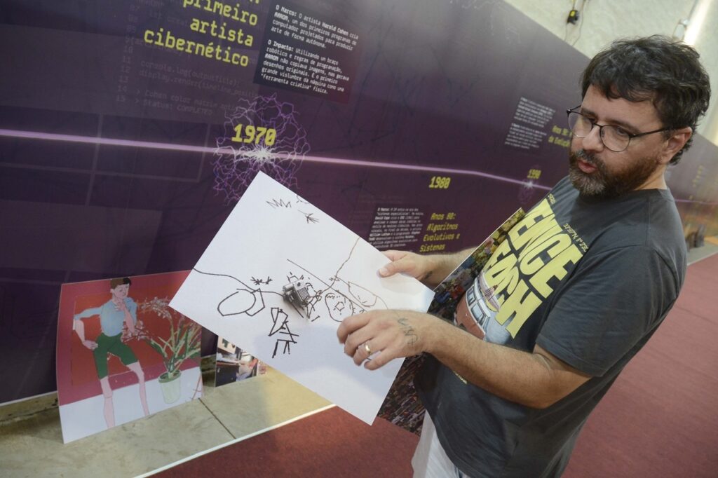 A imagem mostra um homem de óculos e barba, usando uma camiseta escura com letras amarelas, em uma exposição. Ele segura uma folha branca com desenhos feitos à mão e um pequeno dispositivo eletrônico preso ao papel. Ao fundo, há um painel roxo com textos sobre arte cibernética, programação e tecnologia, além de datas como 1970 e 1980. Também aparecem imagens e obras expostas, incluindo uma ilustração colorida apoiada no chão.
