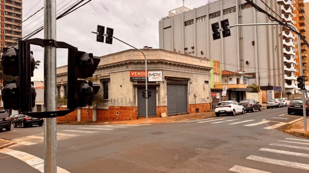 A imagem mostra um cruzamento urbano em uma área central, com faixas de pedestres, semáforos e muitos fios elétricos atravessando o céu. No centro há um prédio antigo, de fachada bege e base de tijolos aparentes, com portas metálicas fechadas e pichações. Uma placa anuncia a venda do imóvel. Ao redor, aparecem edifícios residenciais altos, casas e vários carros estacionados ao longo da rua. O céu está nublado, e a cena tem uma tonalidade levemente alaranjada.