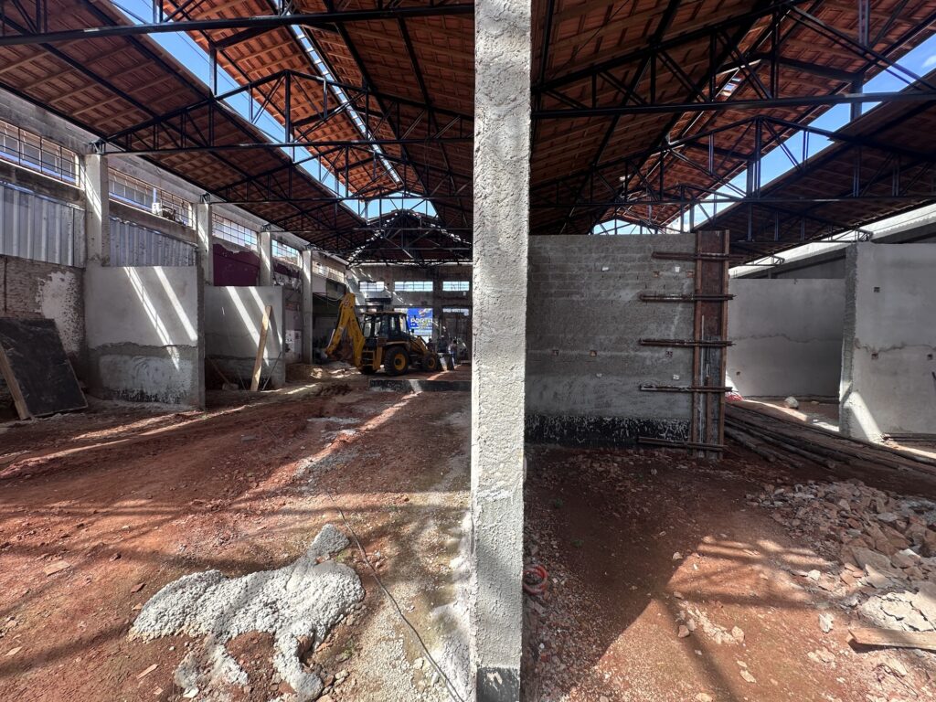 Na fotografia, vemos o interior amplo de uma construção industrial ou comercial em andamento, com o teto elevado sustentado por treliças metálicas pretas e coberto por telhas avermelhadas. O chão ainda é de terra e entulho, com alguns trechos de concreto e restos de obra espalhados. Ao fundo, um trator escavadeira amarelo está estacionado próximo a uma parede parcialmente erguida, onde se destacam tijolos, concreto e barras de reforço expostas, além de andaimes de madeira. A luz natural entra pelas janelas superiores, projetando sombras que reforçam a atmosfera de transformação e progresso estrutural.