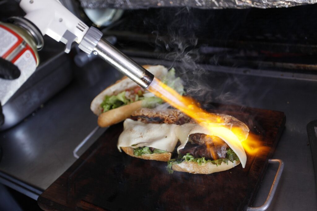 A imagem mostra dois hambúrgueres artesanais sendo finalizados em uma chapa quente. Eles têm pão, hambúrguer de carne, queijo derretido e alface; um maçarico culinário aquece a parte superior, produzindo chamas e bastante vapor. Ao fundo, aparecem utensílios de cozinha e uma embalagem parcialmente visível.
