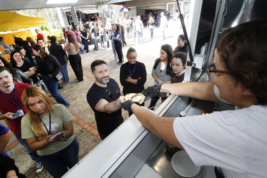 A imagem mostra uma cena movimentada em um evento ao ar livre, onde várias pessoas aguardam em uma fila para serem atendidas em um food truck ou estande de comida. O atendimento: Em primeiro plano, um homem de camiseta preta, com barba e óculos pendurados na gola, está recebendo um prato (que parece uma mini pizza ou lanche aberto) de uma atendente que está dentro do estande, usando luvas pretas de proteção. A fila: Há um grupo de pessoas esperando sua vez. Entre elas, destaca-se uma jovem em primeiro plano, usando uma camiseta verde clara com estampa e segurando um celular, olhando para baixo. Logo atrás do homem que está sendo servido, há uma mulher de blusa preta com um crachá, observando a entrega. O ambiente: O local parece ser um estacionamento ou área pavimentada, com bastante luz do dia. Ao fundo, vê-se outras pessoas circulando, tendas amarelas e estruturas metálicas, indicando que é um evento com diversas opções de alimentação e público presente. O clima parece ser casual e descontraído.