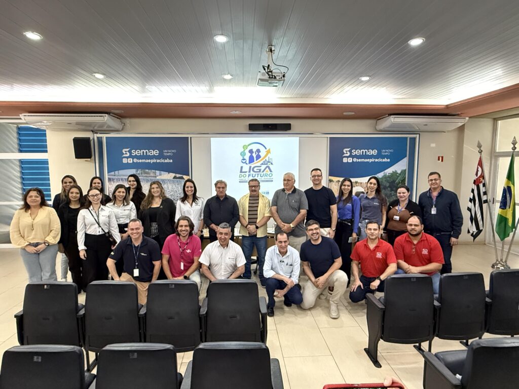A imagem mostra um grupo de cerca de vinte pessoas reunidas em um auditório para uma foto oficial. Ao fundo, há um telão exibindo o logotipo do projeto “Liga do Futuro”, iniciativa da Prefeitura de Piracicaba voltada à orientação profissional e desenvolvimento de carreira. Os participantes estão sorridentes, indicando um clima de colaboração e engajamento. Nas laterais, banners do SEMAE Piracicaba reforçam a parceria institucional, e as bandeiras do Brasil e do Estado de São Paulo completam o cenário.