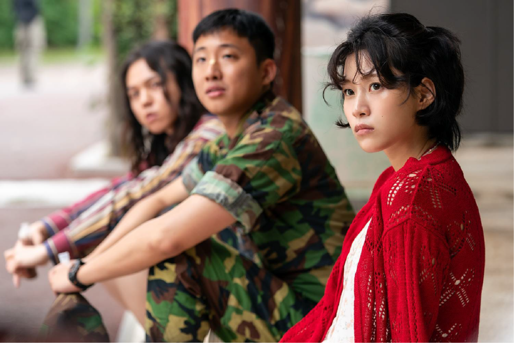 A imagem mostra três jovens sentados lado a lado em um ambiente externo, provavelmente em um momento de pausa ou reflexão. A pessoa em primeiro plano, uma mulher com expressão séria, veste um cardigã vermelho sobre uma blusa clara, destacando-se visualmente. Atrás dela, há um homem com uniforme camuflado e outro com camisa listrada colorida, ambos com olhares voltados para frente.