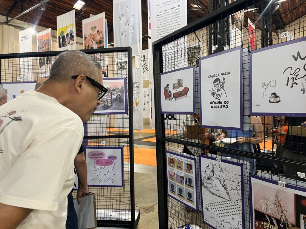 A imagem mostra uma exposição de desenhos e cartuns montada em painéis de grade preta, dentro de um galpão com teto alto de madeira e estrutura metálica. Em primeiro plano, à esquerda, há um homem de cabelos grisalhos, usando óculos escuros e camiseta branca, observando as obras. Os trabalhos estão emoldurados com bordas roxas e identificados por números, como 19, 20, 21, 22 e 23. Um dos cartuns traz o texto “CABEÇA VAZIA — OFICINA DO ALGORITMO”. Ao fundo, há outros desenhos pendurados em grandes faixas, pessoas visitando a exposição e áreas do piso marcadas com faixas laranjas.