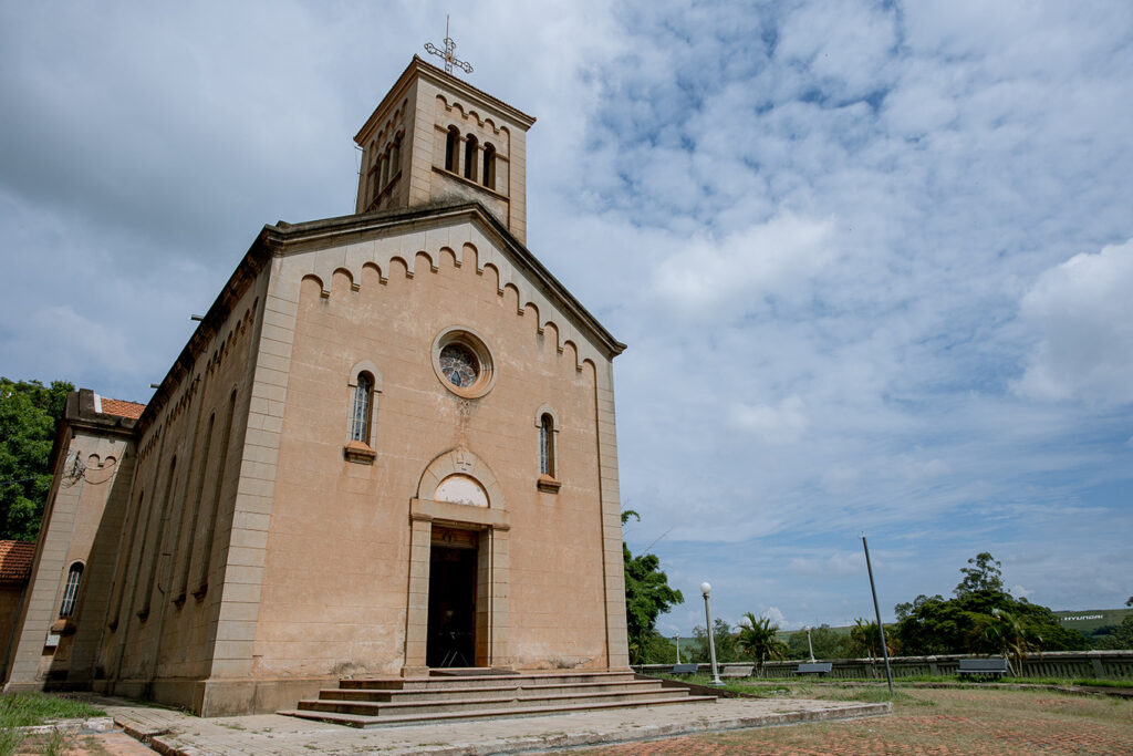 A imagem mostra uma igreja de estilo tradicional, construída em tom bege, com uma torre quadrada encimada por uma cruz metálica. A fachada apresenta janelas em arco, um vitral circular acima da porta principal e detalhes decorativos sob o telhado. O entorno é aberto, com piso de tijolos e vegetação ao fundo, sob um céu parcialmente nublado.