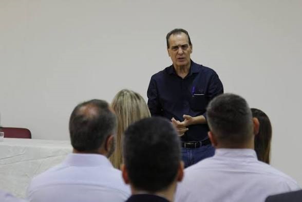 A imagem mostra um pequeno grupo de pessoas participando de uma reunião, palestra ou treinamento em um ambiente interno. Palestrante: Em pé, na frente do grupo, um homem de meia-idade com uma camisa social azul-escura e calça jeans está gesticulando enquanto fala, aparentemente conduzindo a apresentação. Público: Vemos as costas de três pessoas em primeiro plano, sentadas e voltadas para o palestrante. Duas delas estão usando camisas sociais de cor clara. Ambiente: O local possui paredes brancas simples e há uma mesa com toalha branca à esquerda. A iluminação é clara e uniforme, sugerindo um escritório, sala de conferências ou sala de aula.