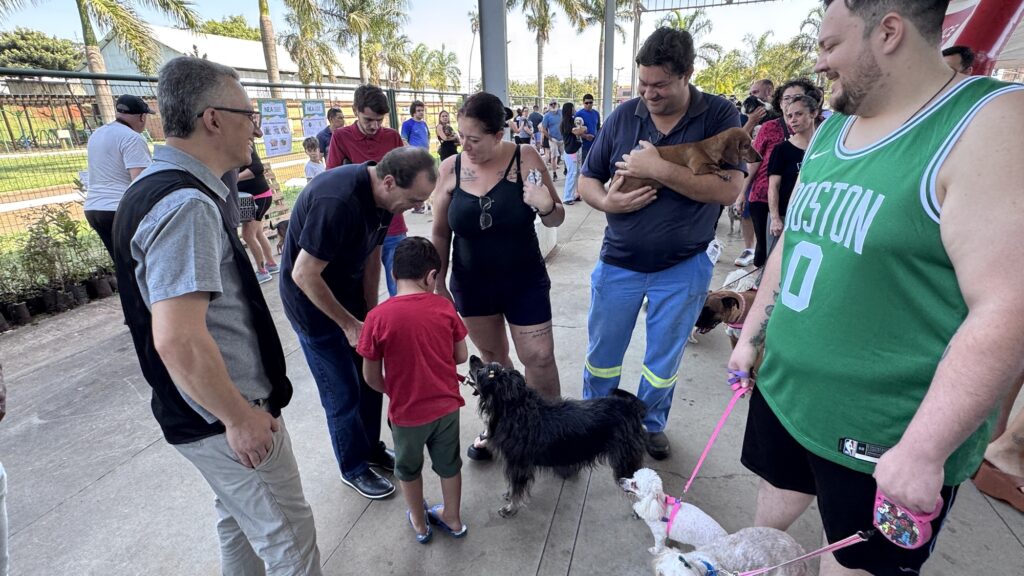 Na imagem, um grupo de pessoas está reunido em uma área pavimentada ao ar livre, próxima a um parque cercado com palmeiras ao fundo. Vários participantes interagem com cães, alguns conduzidos por coleiras e um sendo carregado nos braços. Há cartazes informativos fixados na cerca, reforçando o caráter comunitário do encontro.