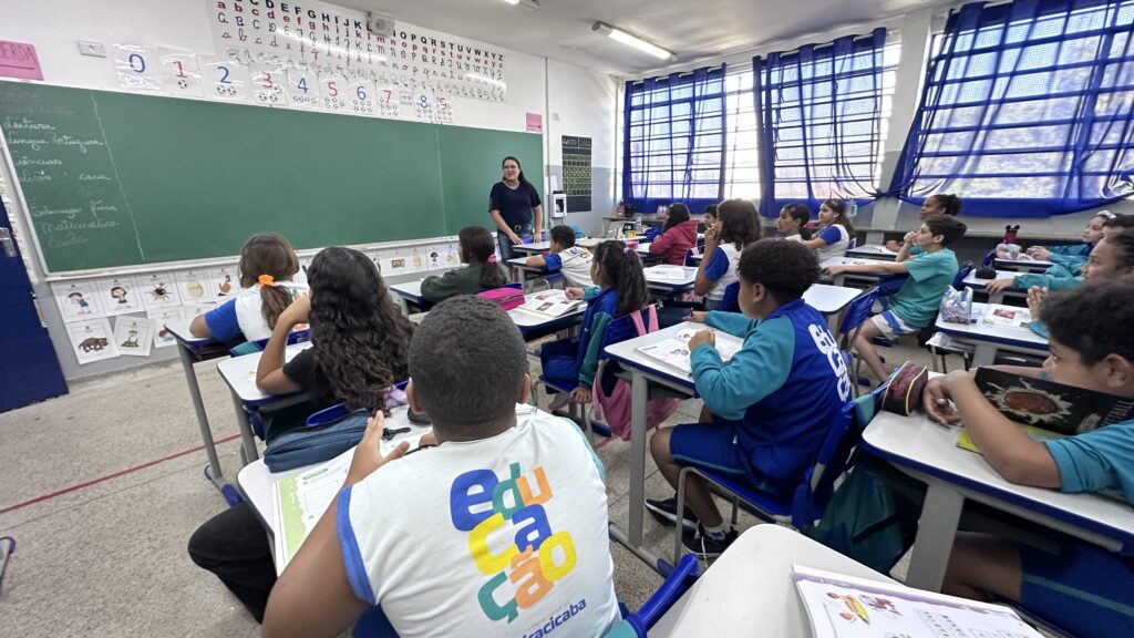 A imagem mostra uma sala de aula cheia de estudantes sentados em carteiras, voltados para a professora, que está em pé diante do quadro. O quadro verde ocupa grande parte da parede e contém algumas anotações escritas com giz. Acima dele, há cartazes educativos com o alfabeto, números, formas de escrita e sinais. À direita, grandes janelas com grades azuis deixam entrar bastante luz. Os alunos usam uniformes coloridos e têm livros, cadernos e estojos sobre as mesas, criando uma cena de aula movimentada e participativa.