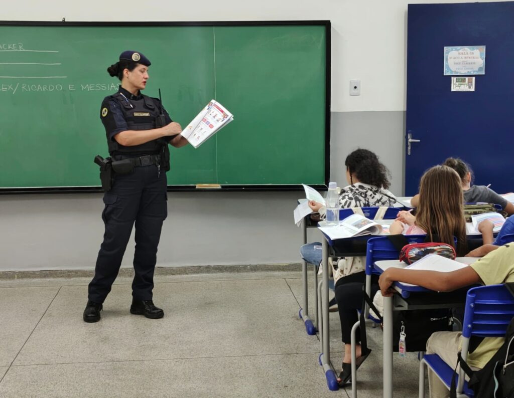 Uma policial em sala de aula ministrando uma atividade para alunos: Pessoa em pé à esquerda: policial fardada (colete tático, coldre, rádio, boina) segurando um caderno/cartilha aberta e apontando/explicando o conteúdo. Quadro-negro verde ao fundo com alguns nomes escritos à esquerda; parede com parte inferior cinza e porta azul à direita. Alunos sentados à direita em carteiras azuis, com cadernos e apostilas abertos; alguns leem enquanto outros têm materiais sobre a mesa (estojo, garrafa de água). Ambiente: sala escolar clara, piso de cerâmica ou granito, atmosfera de aula/encaminhamento educativo. Composição: foco na interação professora–policial e nos estudantes, cena transmite atividade educativa ou palestra sobre segurança/ cidadania.