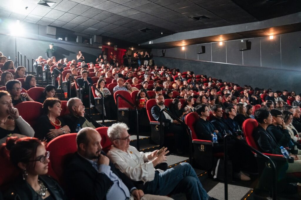 A imagem mostra uma sala de cinema ou auditório cheia de pessoas sentadas em poltronas vermelhas, todas voltadas para a frente, assistindo atentamente a uma apresentação ou exibição. O ambiente é escuro, com iluminação suave nas laterais e teto revestido de painéis acústicos.