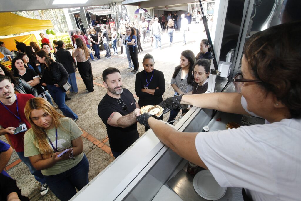 A foto mostra uma movimentada feira ou evento ao ar livre, com várias pessoas reunidas em torno de um food truck. No primeiro plano, uma pessoa usando luvas pretas entrega um prato com um sanduíche ou hambúrguer a um homem de camiseta preta. À direita, uma funcionária de camiseta branca trabalha dentro do veículo. Ao fundo, há filas de pessoas, barracas coloridas — incluindo uma amarela — e outros estandes de comida.