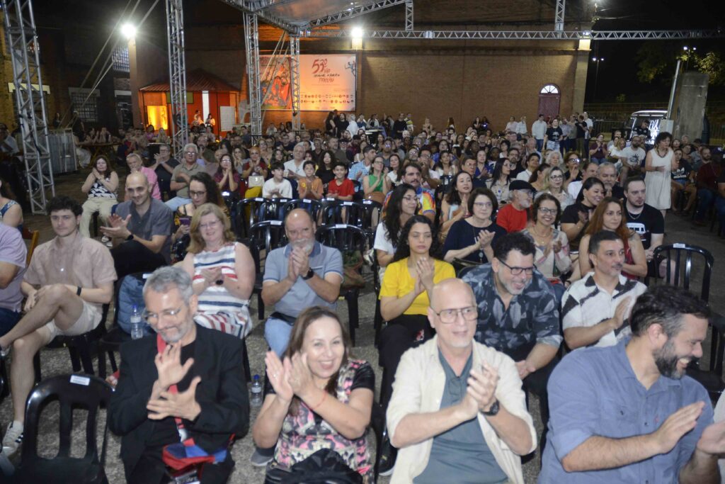A imagem mostra uma grande plateia reunida em um evento noturno ao ar livre. Centenas de pessoas estão sentadas em cadeiras ou em pé, muitas sorrindo, acenando e olhando para a câmera. Ao fundo, há uma estrutura metálica com refletores, um palco parcialmente visível e um grande painel iluminado em tons de laranja. O evento acontece diante de um prédio de tijolos, criando uma atmosfera animada e festiva.