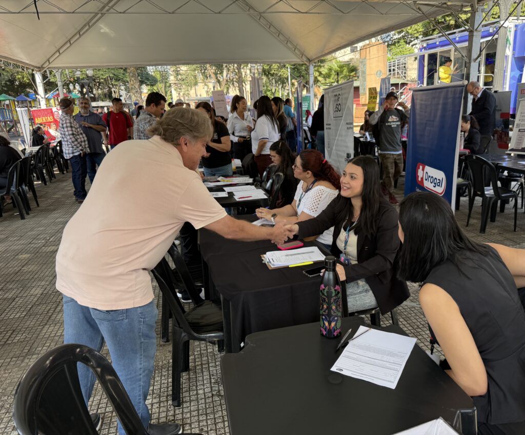 A imagem mostra um evento ao ar livre, realizado sob uma grande tenda branca, com várias mesas cobertas por toalhas pretas e cadeiras de plástico. Pessoas estão interagindo em um ambiente que parece ser uma feira de empregos ou ação de recrutamento profissional.