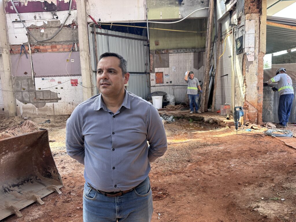 Na cena, uma pessoa de camisa cinza abotoada e jeans está de pé sobre o chão de terra, com as mãos para trás, diante de uma construção parcialmente demolida ou em reforma. Ao fundo, dois trabalhadores com uniformes cinza e faixas refletivas atuam próximos a uma parede de tijolos expostos — um parece coçar a cabeça enquanto o outro trabalha. O ambiente mostra sinais claros de obra, com entulhos, fios, azulejos soltos e equipamentos como um grande balde metálico de máquina pesada e uma furadeira no chão.