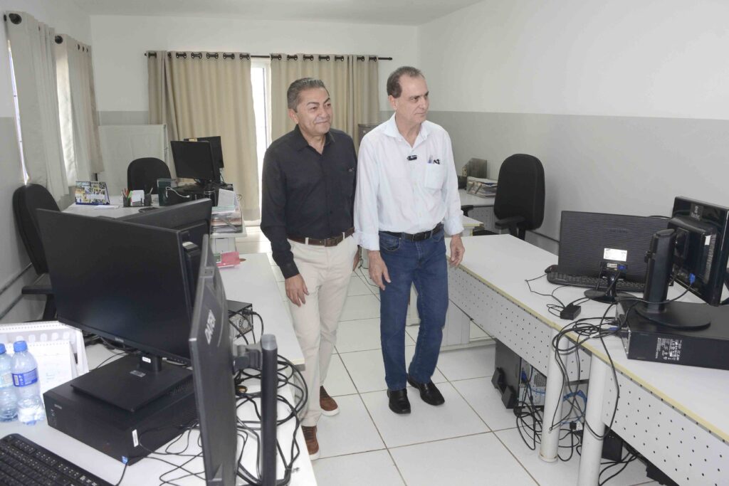 A imagem mostra dois homens em um escritório moderno, caminhando entre mesas equipadas com computadores, monitores e cabos organizados. O ambiente é bem iluminado, com paredes claras, piso branco e cortinas bege, transmitindo uma atmosfera limpa e profissional. Ambos estão vestidos de forma formal — um com camisa preta e calça clara, o outro com camisa branca e jeans — sugerindo uma visita técnica ou inspeção administrativa.