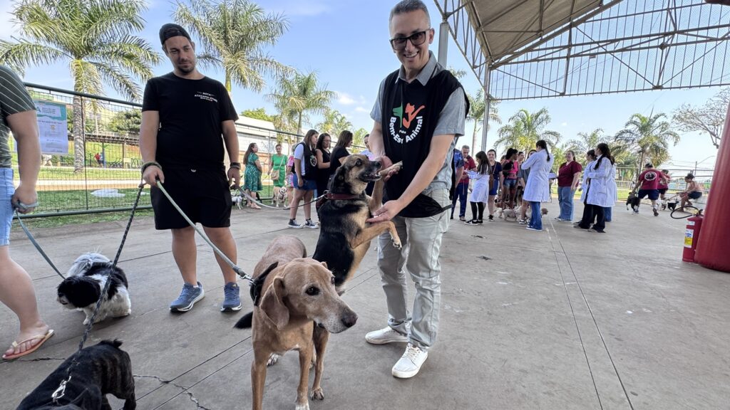 Na imagem, vemos um evento comunitário voltado para o bem-estar animal, realizado em um espaço amplo e coberto. Em primeiro plano, duas pessoas seguram coleiras de vários cães: uma delas interage com um cachorro de porte médio que se apoia nas patas traseiras, enquanto a outra conduz dois cães menores, incluindo um preto e branco. Ao fundo, há mais participantes, alguns com jalecos brancos, sugerindo a presença de veterinários ou voluntários.