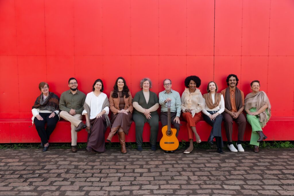 Um grupo de dez pessoas está sentado lado a lado em um banco vermelho diante de uma parede igualmente vermelha, criando um contraste marcante com suas roupas em tons terrosos — marrons, verdes, beges e alaranjados. A cena transmite harmonia visual, equilibrando o fundo vibrante com a paleta natural dos trajes, enquanto um violão apoiado no chão, próximo ao centro, sugere um clima descontraído e talvez musical. O chão de pedras e o toque de verde sob o banco completam a composição, dando à imagem uma atmosfera acolhedora e artística.
