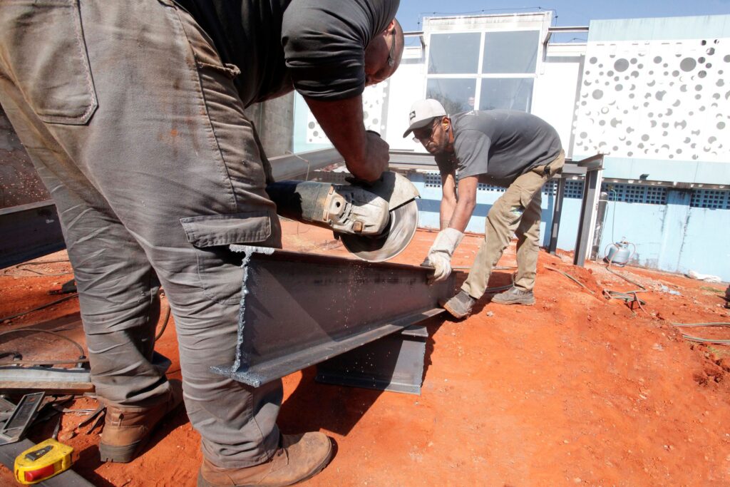 Esta fotografia documental registra o trabalho árduo de dois operários da construção civil utilizando uma esmerilhadeira para cortar uma viga de aço em um canteiro de obras ao ar livre. A composição é estruturada com um foco em profundidade, posicionando o primeiro trabalhador em primeiro plano, quase inteiramente enquadrado, enquanto o segundo operário atua como contraponto visual no plano intermediário. O ângulo de câmera, posicionado ligeiramente acima, permite observar a interação direta entre a ferramenta e o material metálico. O movimento é capturado no momento exato do corte, com o operário em primeiro plano aplicando força enquanto a esmerilhadeira gera faíscas. A organização espacial enfatiza a colaboração, com um homem segurando o material e o outro operando o equipamento, criando uma narrativa visual de esforço técnico e precisão. O primeiro operário, visto de costas, veste uma camisa preta e calças de sarja cinza com bolsos utilitários, calçando botas de trabalho robustas. Seus braços estão tensionados pelo esforço de manusear a máquina pesada. O segundo trabalhador, posicionado de frente para o espectador, usa uma camiseta cinza, um boné claro e calças cáqui. Ele usa luvas de proteção cinzas e mantém uma postura de vigilância, segurando a viga para garantir a estabilidade do corte. Suas feições demonstram concentração e foco na tarefa, refletindo o profissionalismo necessário para operar ferramentas de alto risco em um ambiente de construção. A imagem é uma fotografia de estilo realista e técnico, caracterizada por um registro nítido e uma profundidade de campo que mantém tanto a ação próxima quanto o fundo em foco razoável. A execução técnica privilegia a clareza, utilizando uma velocidade de obturador adequada para congelar as faíscas que emanam do ponto de contato entre a esmerilhadeira e a viga metálica. A paleta de cores é dominada pelos tons terrosos do solo, que contrastam fortemente com o cinza frio das vigas metálicas e o vestuário dos trabalhadores, criando uma distinção visual clara entre o ambiente de terra vermelha e os materiais de construção industrial. O cenário é um canteiro de obras aberto, caracterizado por um solo de terra batida de cor avermelhada vibrante, típico de regiões onde o solo é rico em ferro. Ao fundo, vislumbra-se uma estrutura arquitetônica moderna com paredes azuis e janelas amplas, composta por padrões geométricos em relevo que contrastam com a crueza do local de trabalho. A iluminação é de luz natural direta, sugerindo um dia claro com céu aberto, o que intensifica o contraste e projeta sombras definidas sob a viga. O ambiente transmite uma atmosfera de atividade industrial intensa e produtividade, onde elementos simples como uma trena amarela no canto inferior esquerdo reforçam a natureza prática e cotidiana da cena.