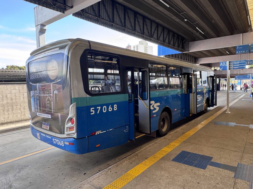 A imagem mostra um ônibus urbano azul e prata estacionado em um terminal coberto, identificado pelo número 57068. Na parte traseira, há um anúncio do O Boticário promovendo o Dia dos Pais, com produtos de perfumaria. O local tem sinalização azul indicando plataformas e linhas, além de piso tátil amarelo e azul para acessibilidade. Algumas pessoas aparecem ao fundo, e a estrutura do terminal é feita de concreto e metal, típica de áreas de transporte público em grandes cidades.