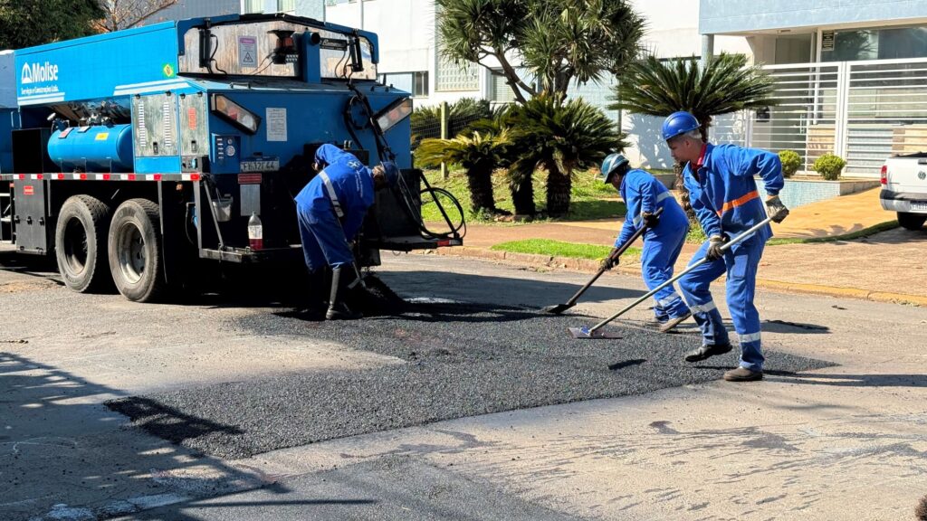 Você tem razão — olhando com atenção, os capacetes dos trabalhadores são azuis, não brancos. Na cena, três homens com uniformes azuis e capacetes da mesma cor trabalham juntos na manutenção de uma rua residencial, espalhando e nivelando o asfalto preto recém-saído de um caminhão azul da empresa “Molise Serviços e Construções Ltda.”; ao redor, casas em tons claros, plantas verdes e uma caminhonete branca estacionada completam o cenário.
