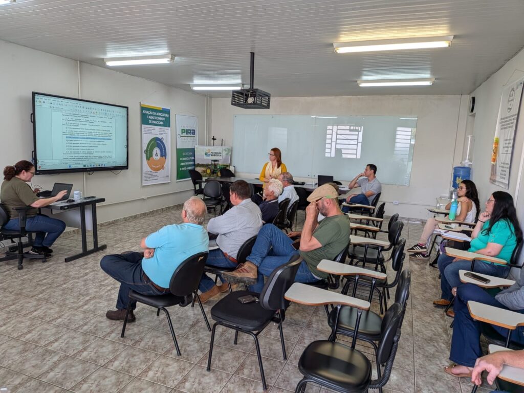 Sala de reunião/treinamento com apresentação em andamento. Pessoas: cerca de 12 participantes adultos sentados em cadeiras com prancheta; alguns prestam atenção, outros conversam ou olham para o celular. Apresentação: uma mulher em pé (cardigã amarelo) parece estar falando; outra pessoa sentada à esquerda opera um notebook ligado a uma grande tela na parede que exibe um documento/arquivo de texto. Ambiente: piso de cerâmica com padrões, teto com luminárias fluorescentes e projetor suspenso no centro. Mobiliário: cadeiras tipo sala de aula com prancheta acoplada, mesa de apresentador à esquerda, e uma grande tela/monitor fixo na parede. Materiais e identificação: banners na parede com logotipos e textos sobre “ATUAÇÃO DA AGRICULTURA E ABASTECIMENTO DE PIRACICABA” e um display com o logotipo “PIRA”. Há também uma garrafa d’água grande em bebedouro à direita. Iluminação e janelas: luz artificial predominante; há janelas ao fundo que deixam entrar luz natural difusa. Atmosfera: reunião técnica ou institucional, tom sério e funcional, foco em apresentação de documento/informações.