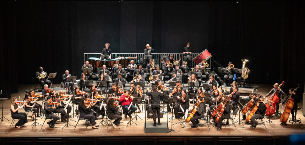 A orquestra completa, com pelo menos cinquenta músicos, está apresentada em um palco de madeira, com um fundo preto uniforme. A orquestra está disposta em várias fileiras em semicírculo, com os músicos mais altos e os instrumentos de percussão posicionados nas fileiras traseiras e mais elevadas. A iluminação é focada no palco, destacando os músicos e seus instrumentos. A paleta de cores predominante é composta por tons escuros, como o preto do fundo e do vestuário de muitos músicos, contrastando com a madeira clara do palco e o brilho metálico dos instrumentos de sopro e de corda. Na fileira da frente, um maestro, vestindo um terno preto, está de costas para a câmera, de frente para a orquestra, com os braços erguidos, parecendo reger a performance. Ao lado dele, estão músicos tocando violoncelos e contrabaixos. A maioria dos músicos nas fileiras intermediárias e frontais está tocando violinos, violas e instrumentos de sopro como clarinetes e flautas. Nas fileiras mais altas e traseiras, observamos músicos tocando instrumentos de sopro de metal, como trombetas, trombones e tubas, além de percussionistas com tímpanos e um tambor vermelho proeminente. Alguns músicos estão sentados em cadeiras, enquanto outros parecem estar em assentos elevados. Um músico está sentado em uma cadeira de rodas, tocando um instrumento. A composição da imagem é panorâmica, capturando a totalidade da orquestra e do palco. A profundidade é criada pela disposição em fileiras dos músicos e pela iluminação. Detalhes notáveis incluem as partituras nos atrís à frente de cada músico e a variedade de instrumentos que compõem a orquestra. A imagem transmite uma atmosfera de profissionalismo e dedicação musical.