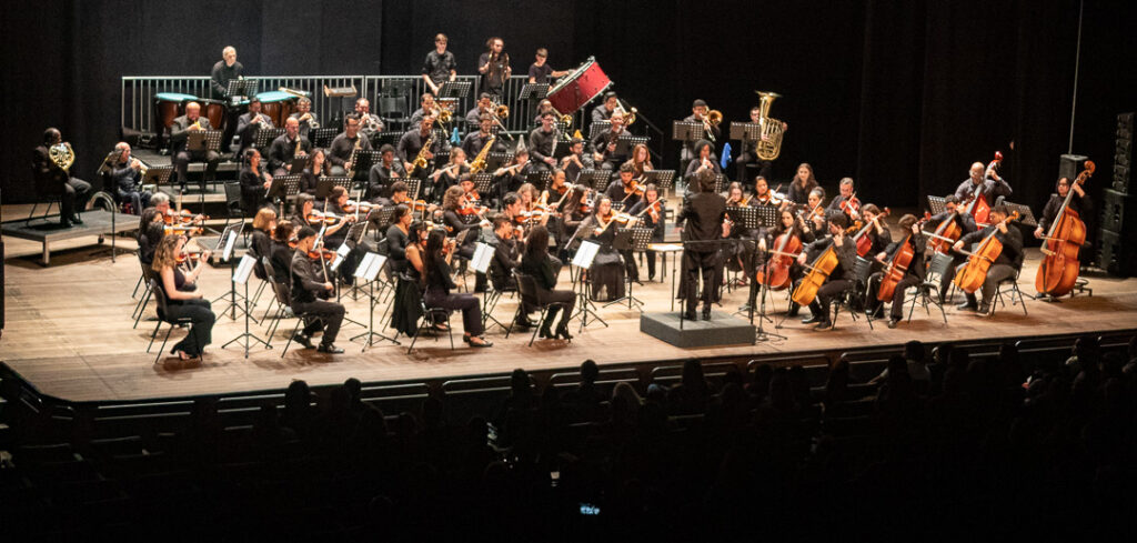 A orquestra completa, com pelo menos cinquenta músicos, está apresentada em um palco de madeira, com um fundo preto uniforme. A orquestra está disposta em várias fileiras em semicírculo, com os músicos mais altos e os instrumentos de percussão posicionados nas fileiras traseiras e mais elevadas. A iluminação é focada no palco, destacando os músicos e seus instrumentos. A paleta de cores predominante é composta por tons escuros, como o preto do fundo e do vestuário de muitos músicos, contrastando com a madeira clara do palco e o brilho metálico dos instrumentos de sopro e de corda. Na fileira da frente, um maestro, vestindo um terno preto, está de costas para a câmera, de frente para a orquestra, com os braços erguidos, parecendo reger a performance. Ao lado dele, estão músicos tocando violoncelos e contrabaixos. A maioria dos músicos nas fileiras intermediárias e frontais está tocando violinos, violas e instrumentos de sopro como clarinetes e flautas. Nas fileiras mais altas e traseiras, observamos músicos tocando instrumentos de sopro de metal, como trombetas, trombones e tubas, além de percussionistas com tímpanos e um tambor vermelho proeminente. Alguns músicos estão sentados em cadeiras, enquanto outros parecem estar em assentos elevados. Um músico está sentado em uma cadeira de rodas, tocando um instrumento. A composição da imagem é panorâmica, capturando a totalidade da orquestra e do palco. A profundidade é criada pela disposição em fileiras dos músicos e pela iluminação. Detalhes notáveis incluem as partituras nos atrís à frente de cada músico e a variedade de instrumentos que compõem a orquestra. A imagem transmite uma atmosfera de profissionalismo e dedicação musical.