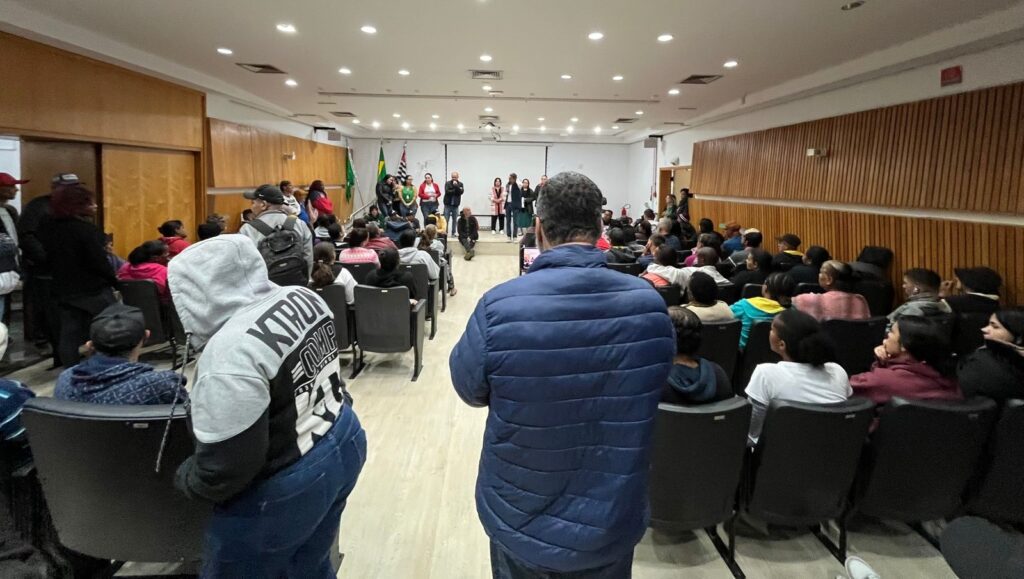 Um grupo de bolsistas participa de uma reunião em um auditório, onde recebe orientações de representantes sobre a inscrição para uma prova.