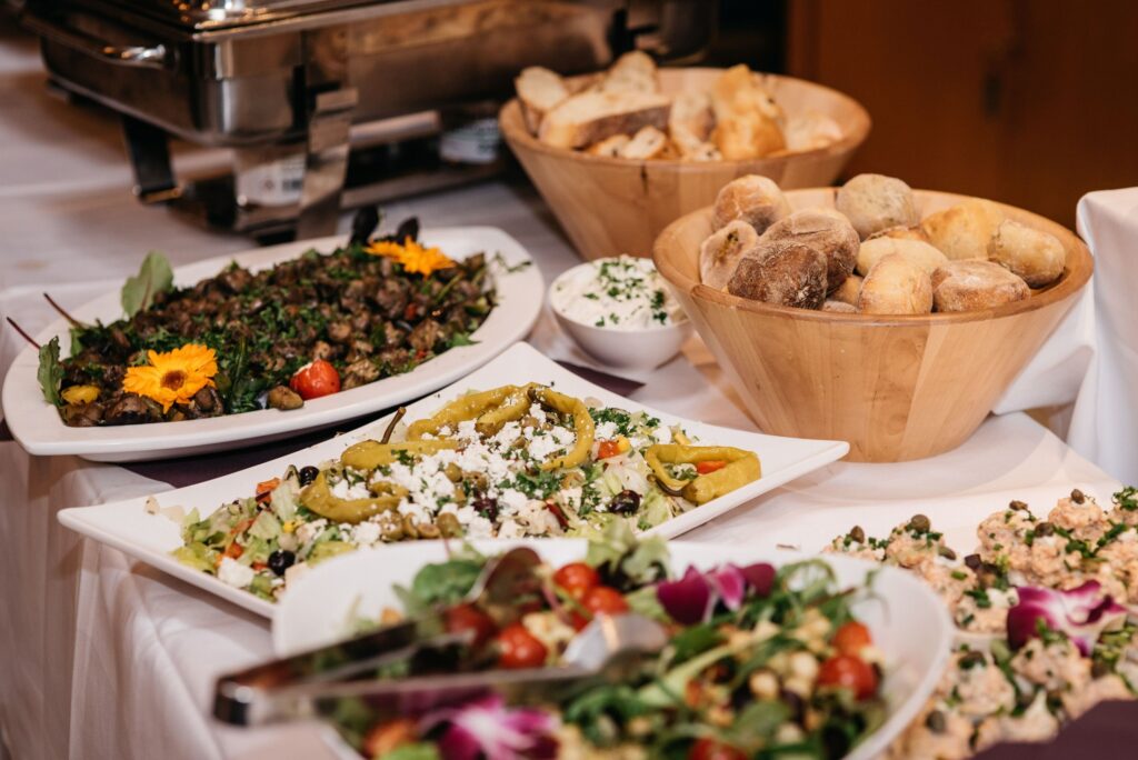 A imagem mostra uma mesa de buffet bem servida, coberta com toalha branca. Há várias travessas com saladas, legumes, azeitonas, queijo branco e outros acompanhamentos decorados com ervas e flores comestíveis. À direita, aparecem cestos de madeira cheios de pães variados. Ao fundo, há equipamentos metálicos de cozinha e recipientes com mais alimentos. A iluminação quente e os pratos coloridos dão à cena um aspecto elegante e convidativo, típico de um evento ou restaurante.