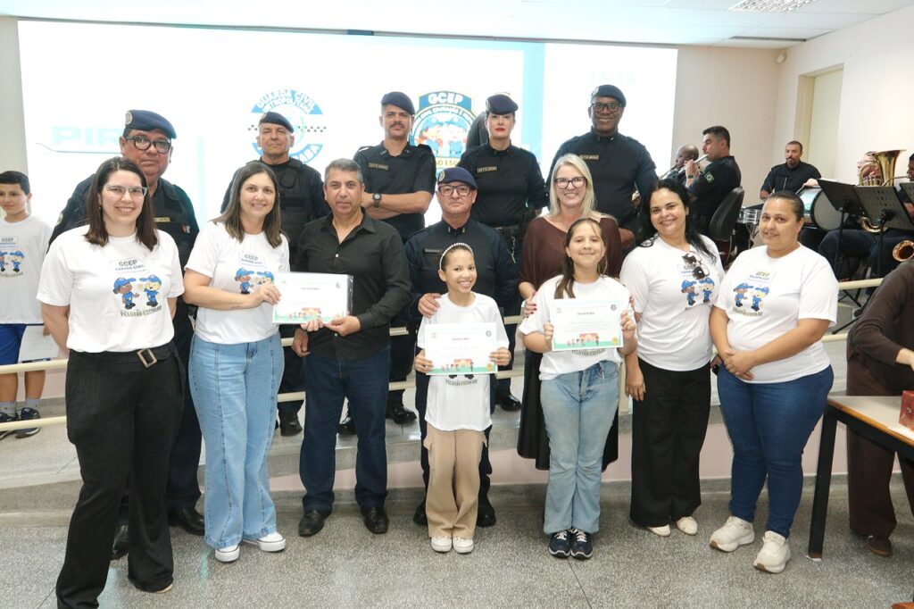 A imagem registra um momento de entrega de certificados durante a cerimônia de formatura do GCEP. Em primeiro plano, duas estudantes aparecem segurando seus certificados de conclusão, acompanhadas por familiares, representantes da escola e agentes da Guarda Civil.
Ao fundo, integrantes da Guarda Civil Municipal posam ao lado dos participantes. No telão, aparecem as identidades visuais da Prefeitura de Piracicaba, da Guarda Civil e do GCEP. Também é possível observar músicos da corporação à direita da imagem, com instrumentos musicais.