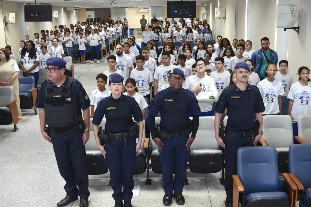 A imagem mostra quatro agentes da Guarda Civil Municipal em primeiro plano, posicionados diante de um auditório. Eles aparecem em pé, lado a lado, durante a cerimônia do GCEP.
Ao fundo, há dezenas de estudantes usando camisetas brancas do programa, além de professores, familiares e outros participantes. Os alunos estão organizados nas fileiras do auditório, acompanhando o momento da cerimônia. A fotografia evidencia a presença dos guardas educadores e a grande participação dos estudantes no evento.