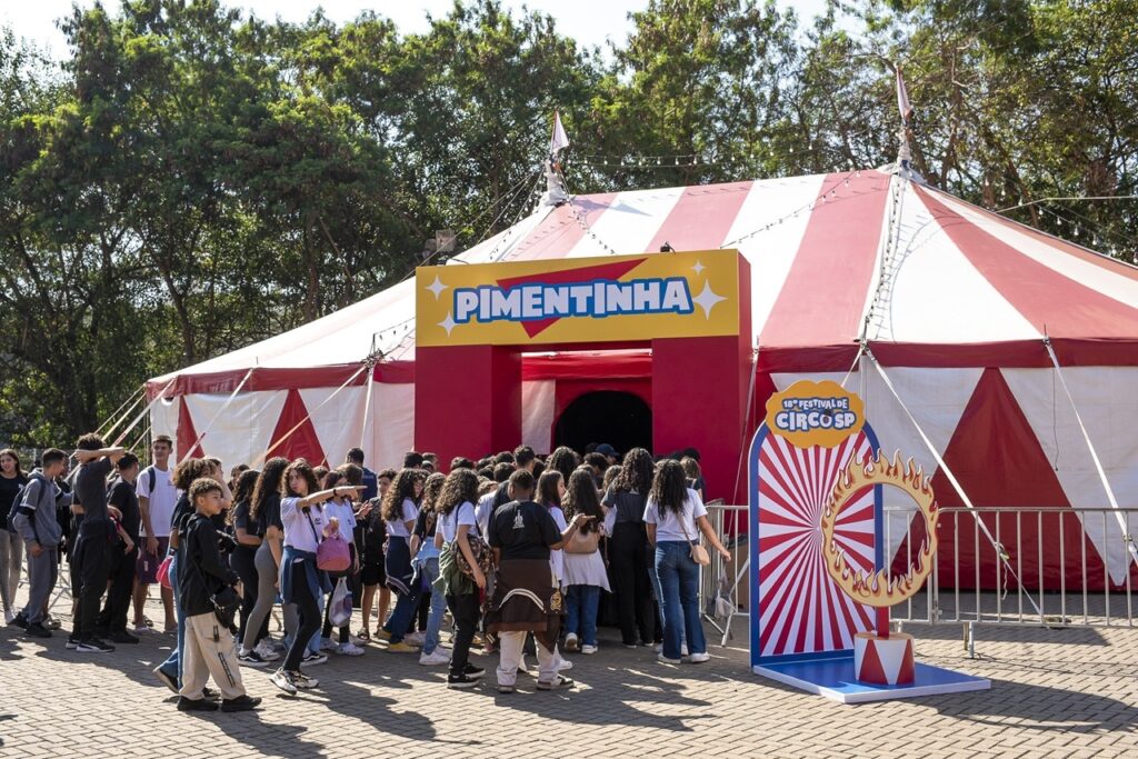 A imagem mostra uma grande tenda de circo listrada em vermelho e branco, instalada em uma área arborizada e ensolarada. Na entrada, há uma placa colorida com o nome “Pimentinha”. À direita, um painel anuncia o 18º Festival de Circo, com uma ilustração de pipoca e um arco em chamas. Em frente à tenda, um grupo numeroso de jovens aguarda para entrar, enquanto grades metálicas organizam a fila.
