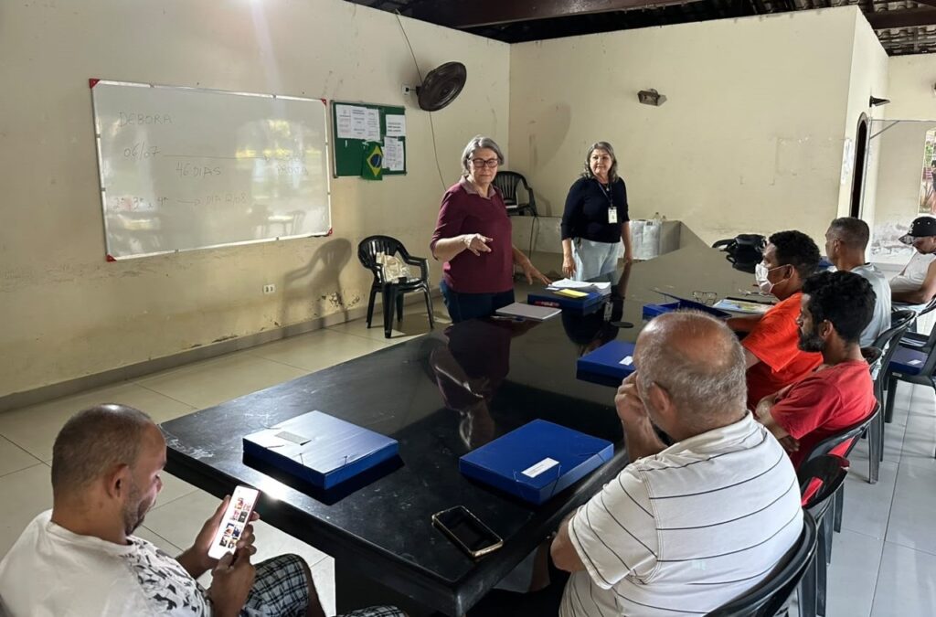 Um grupo de pessoas participa de uma aula de reforço em uma sala, acompanhando as orientações de duas instrutoras. Os participantes estão sentados ao redor de uma mesa, com materiais de estudo.