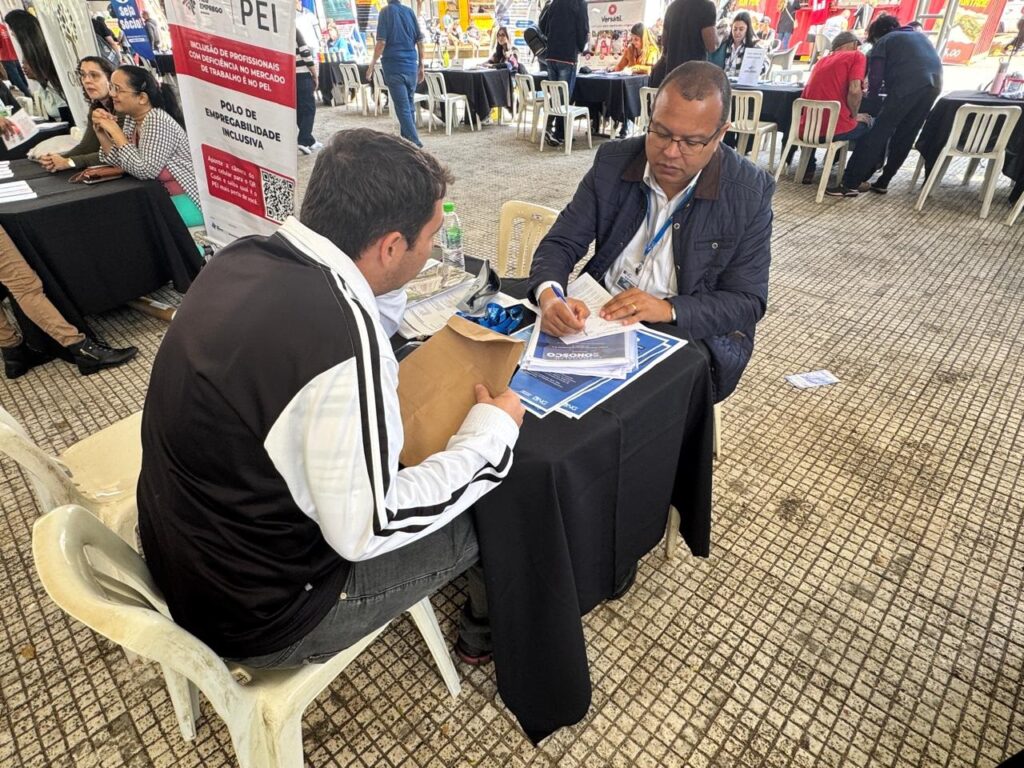 A imagem mostra um momento de atendimento em um evento profissional, provavelmente uma feira de empregabilidade ou feira de empregos. Aqui estão os detalhes principais: A Cena: Dois homens estão sentados frente a frente em uma mesa coberta com uma toalha preta. O homem à direita, usando óculos e uma jaqueta escura, parece estar preenchendo ou revisando documentos. O homem à esquerda, de costas para a câmera e vestindo uma blusa esportiva preta e branca, segura um envelope pardo, possivelmente contendo um currículo. O Ambiente: É um ambiente ao ar livre, mas coberto por uma grande tenda. O chão é pavimentado com blocos de concreto. Há várias outras pessoas ao fundo, sentadas em outras mesas, o que reforça a ideia de um evento com múltiplos estandes de atendimento. Contexto: Ao lado da mesa, há um banner informativo do PEI (Polo de Empregabilidade Inclusiva), que menciona "Inclusão de profissionais com deficiência no mercado de trabalho". O banner também convida o público a usar o celular para acessar um QR Code. Clima: O ambiente é de trabalho e atendimento ao público, com uma organização voltada para o suporte profissional.