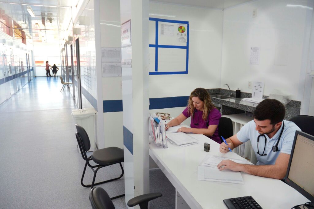 Uma cena serena em um ambiente clínico mostra dois profissionais de saúde focados em suas tarefas administrativas em uma estação de trabalho organizada, enquanto um corredor estendido se estende ao fundo. A composição é dividida em duas áreas distintas: uma estação de trabalho em primeiro plano onde dois profissionais estão sentados lado a lado, e um longo corredor à esquerda que conduz o olhar para o fundo da imagem. O espaço de trabalho é definido por uma bancada branca, onde os profissionais realizam anotações em documentos. A disposição sugere uma rotina funcional de atendimento médico, com cadeiras ergonomicamente posicionadas e uma organização limpa que reflete a natureza profissional do ambiente. Os protagonistas são um homem e uma mulher, ambos vestidos com trajes de trabalho da área da saúde. A mulher, à esquerda, veste uma blusa roxa e tem cabelos claros ondulados; ela está concentrada em escrever em papéis. Ao lado dela, um homem jovem com cabelo escuro curto veste uma camisa polo azul clara e usa um estetoscópio ao redor do pescoço, também concentrado em suas anotações. Suas posturas são de trabalho focado e diligente, com gestos naturais de quem manipula prontuários e materiais administrativos. A imagem é uma fotografia digital que busca registrar a realidade de um ambiente de trabalho hospitalar ou clínico. A execução é direta, sem artifícios estilísticos, priorizando a nitidez e o registro fiel das condições do local. A iluminação é difusa e uniforme, típica de interiores com lâmpadas fluorescentes, o que cria um ambiente claro, higiênico e desprovido de sombras marcantes. A paleta de cores é composta por tons neutros de branco e cinza, contrastando com as vestimentas coloridas dos profissionais, o que ajuda a destacar os sujeitos centrais em meio à simplicidade do consultório. O cenário é um ambiente clínico moderno e funcional, caracterizado por paredes brancas, superfícies de granito e piso liso, transmitindo uma atmosfera de eficiência e seriedade. O corredor lateral apresenta uma iluminação que vem de uma grande abertura ou janela ao fundo, criando um ponto de fuga luminoso. Elementos como uma pequena pia, documentos organizados e equipamentos de escritório completam o cenário, reforçando o caráter prático do ambiente. A atmosfera é de tranquilidade e disciplina, típica de momentos de pausa ou preenchimento de registros em uma unidade de saúde.
