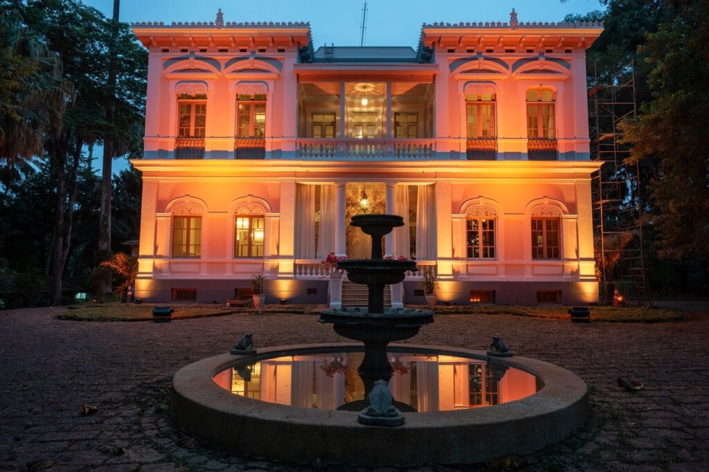 Uma mansão histórica iluminada à noite, vista de frente com uma fonte circular em primeiro plano que reflete a fachada. Arquitetura: prédio de dois andares com ornamentações clássicas — arcos decorados sobre as janelas, cornijas trabalhadas no beiral e sacada central no segundo piso com balaustrada. Iluminação: luzes quentes (tons alaranjados) banham a fachada, realçando detalhes e criando contraste com o céu azul-escuro do crepúsculo. Entrada: porteiro/vestíbulo central recuado, entre duas colunas discretas; cortinas claras nas aberturas laterais do térreo e um lustre ou luminária visível no interior. Fonte: fonte de múltiplos andares diretamente alinhada com a entrada; a lâmina d’água na bacia reflete a luz da fachada; pequenas esculturas de sapo ou similar adornam a borda da bacia. Ambiente externo: calçamento em pedras na frente, canteiros baixos e vasos; árvores altas e vegetação densa cercam a propriedade, criando um enquadramento verde; há andaimes ou estrutura metálica visível no lado direito, indicando obra ou manutenção. Atmosfera: elegante e acolhedora, com sensação de evento ou iluminação cênica que destaca o caráter histórico do imóvel.