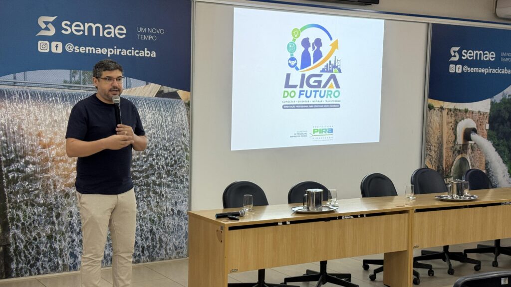 A imagem mostra um homem falando ao microfone durante uma apresentação em um auditório. No fundo, há um telão exibindo o logotipo do projeto “Liga do Futuro”, iniciativa da Prefeitura de Piracicaba voltada à orientação profissional e desenvolvimento de carreira. O ambiente é institucional, com mesas, cadeiras e banners do SEMAE Piracicaba, empresa municipal de saneamento, indicando parceria no evento.