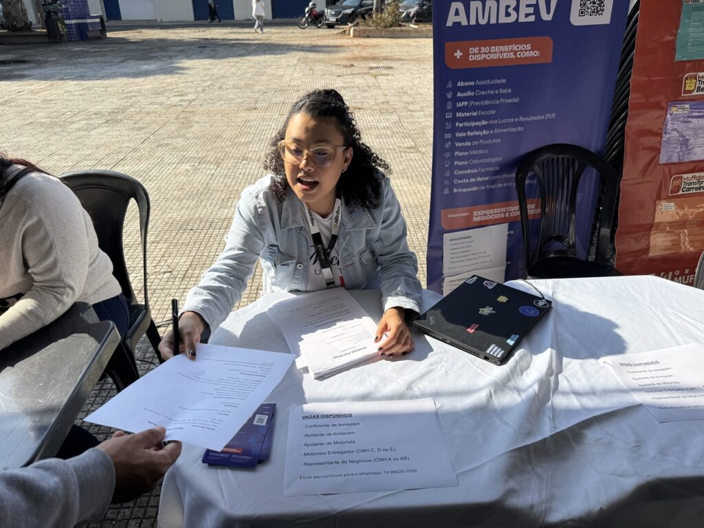 A imagem mostra uma mulher sentada em uma mesa ao ar livre, participando de um evento de recrutamento profissional. Ela entrega uma folha de papel a outra pessoa, enquanto sobre a mesa há documentos, um laptop com adesivos e panfletos com informações sobre vagas de emprego.