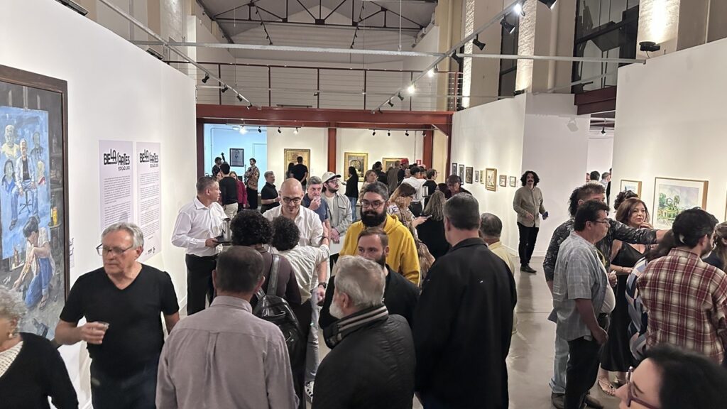 A imagem mostra o vernissage ou a abertura de uma exposição de arte em um espaço amplo e industrial. O Público: O local está movimentado, com várias pessoas circulando, conversando e observando as obras. Vemos visitantes de diversas idades, muitos vestidos de forma casual ou casual-chique. Há pessoas em grupos conversando, outras de costas analisando os quadros nas paredes, e um senhor de camisa branca carregando uma bandeja, sugerindo que há serviço de bebidas ou aperitivos. O Ambiente: O espaço tem um estilo "galpão industrial", com vigas metálicas aparentes, teto alto e paredes brancas que destacam as obras. A iluminação é composta por trilhos de luz direcionados para os quadros. As Obras: As paredes estão repletas de quadros de diferentes tamanhos e estilos. À esquerda, há uma pintura de grande porte que retrata cenas de um ateliê ou ambiente de trabalho. Também há cartazes informativos na parede com o título "Bela Artes - Edição XXXI". Atmosfera: O clima é de um evento cultural social, onde o foco é a interação entre os convidados e a apreciação da exposição de arte.