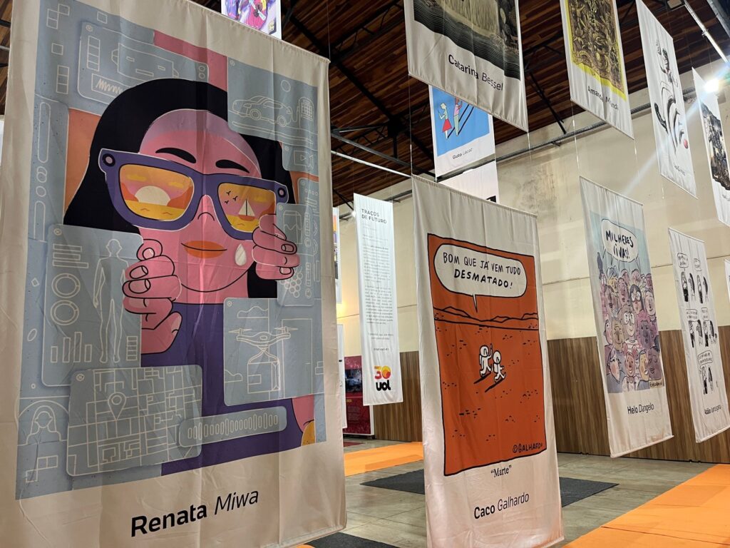 A imagem mostra uma exposição de ilustrações e quadrinhos em um amplo espaço cultural, com obras impressas em grandes painéis de tecido suspensos no teto. Em primeiro plano, há uma ilustração colorida de uma pessoa usando óculos escuros, assinada por Renata Miwa. Ao lado, aparece uma tirinha em tons de laranja, com a frase “Bom que já vem tudo desmatado!”, de Caco Galhardo. Também são visíveis obras de outros artistas, como Catarina Bessell, Guto Lacaz, Amaral, Helo D’Angelo e Arnaldo Branco. Um dos painéis traz o título “Traços de Futuro” e o logotipo dos 30 anos da UOL. Os trabalhos abordam temas sociais, ambientais e cotidianos, com estilos variados, incluindo humor, crítica e arte digital.