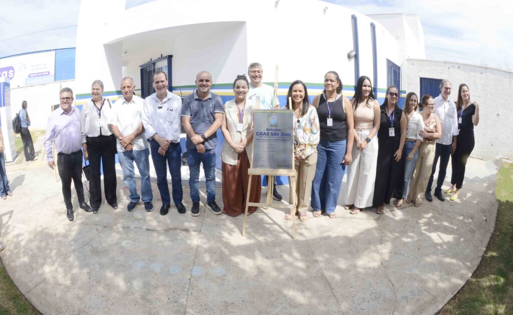descreva brevemente A imagem mostra um grupo de pessoas reunidas em frente a um prédio branco com detalhes em azul e verde, participando de um evento oficial. No centro, há uma placa de inauguração com o título “Reforma CRAS São José”, indicando a reinauguração ou reforma de um Centro de Referência de Assistência Social. As pessoas estão sorridentes, vestidas de forma profissional, e parecem ser autoridades e funcionários envolvidos no projeto. O ambiente é claro e ensolarado, transmitindo um clima de celebração e formalidade.